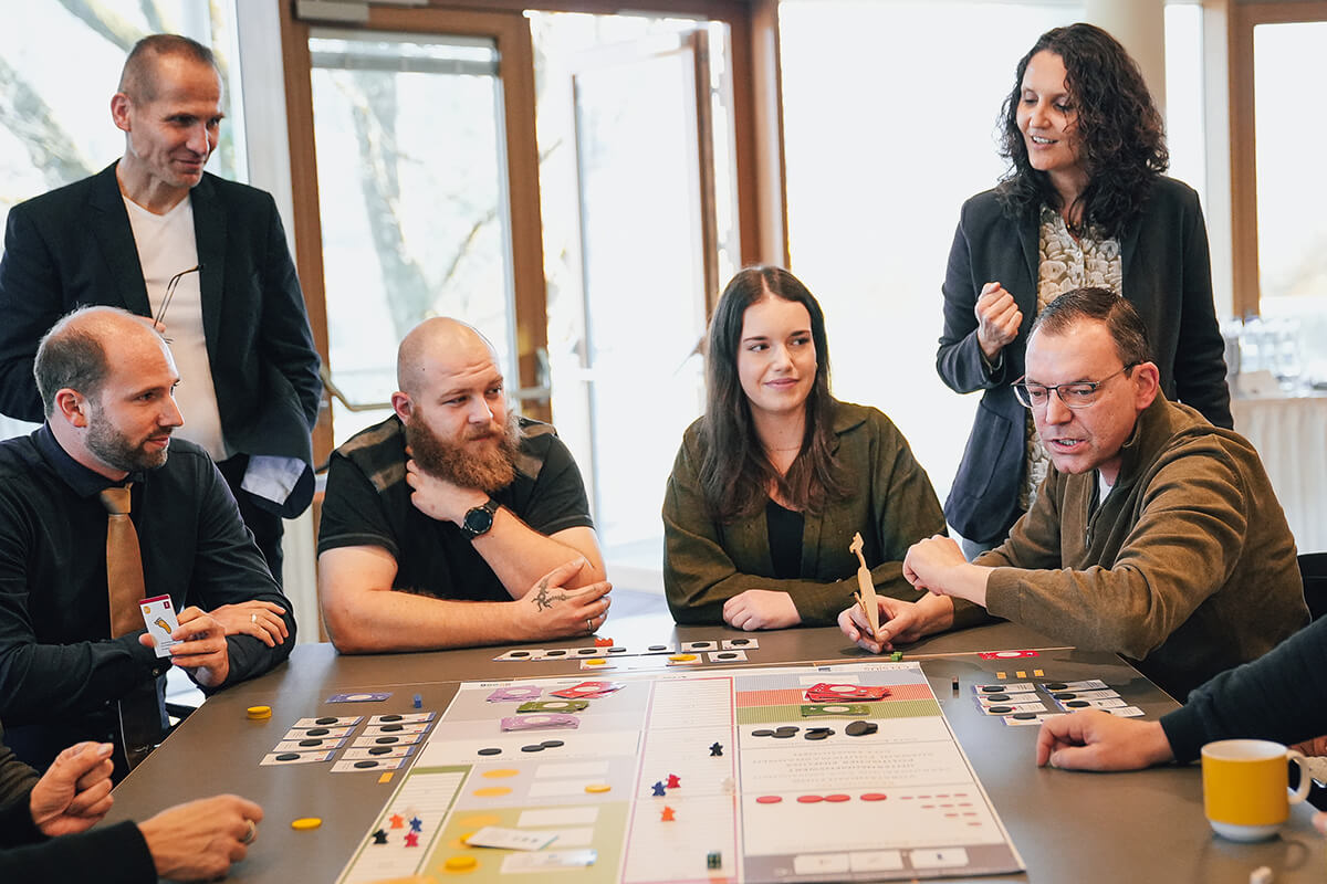 Gruppe im Celsius-Workshop am Spieltisch Mehrere Teilnehmende sitzen und stehen um einen Tisch und spielen Celsius - Die 2 Grad Challenge